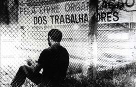 Homem sentado ao lado de faixa onde se lê “Pela livre organização dos Trabalhadores” ([Santo André-SP?], data desconhecida). / Crédito: Roberto Parizotti.